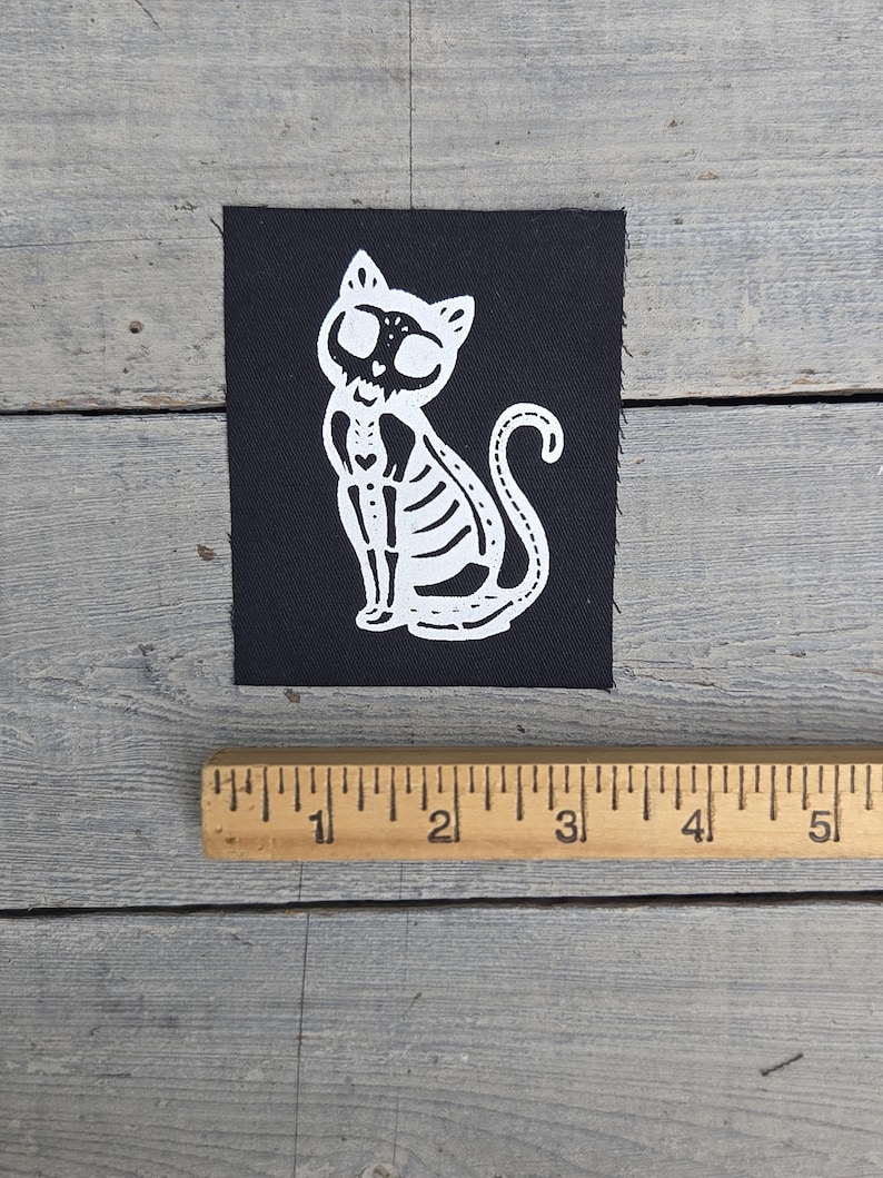 May include: A black square patch with a white cat skeleton design. The cat is sitting upright with its tail curled. The patch is on a grey wooden surface next to a wooden ruler.