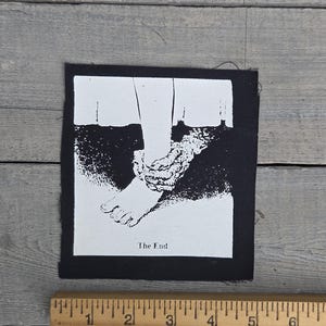 以下が含まれることがあります： 足が縛られたグラフィックイラストが描かれた白黒のパッチ。画像の下には「The End」の文字が印刷されています。パッチは長方形で、幅は約7.6cmです。