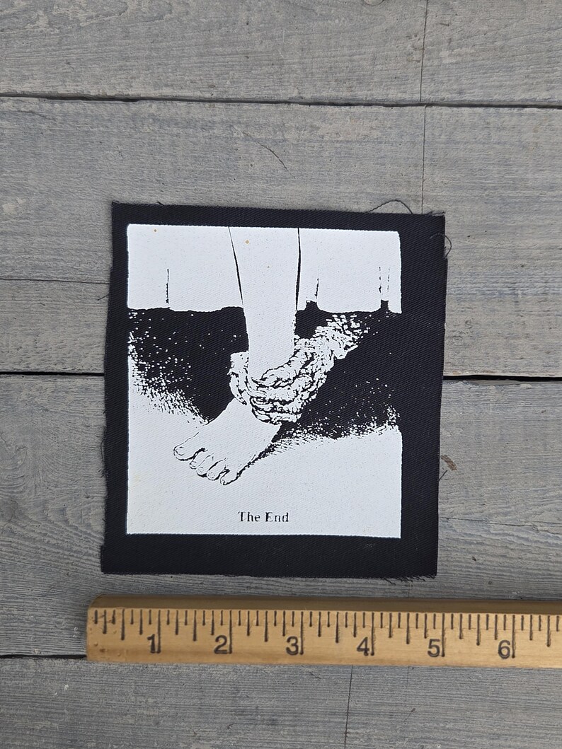 以下が含まれることがあります： 足が縛られたグラフィックイラストが描かれた白黒のパッチ。画像の下には「The End」の文字が印刷されています。パッチは長方形で、幅は約7.6cmです。