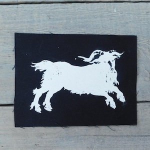 Op de afbeelding: Een zwarte rechthoekige patch met een witte illustratie van een geit in volle sprong. De geit heeft grote, gebogen hoorns en een ruige baard. De patch heeft een ruwe, onafgewerkte rand.