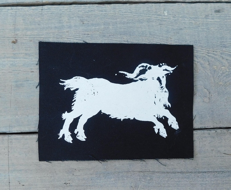 Op de afbeelding: Een zwarte rechthoekige patch met een witte illustratie van een geit in volle sprong. De geit heeft grote, gebogen hoorns en een ruige baard. De patch heeft een ruwe, onafgewerkte rand.