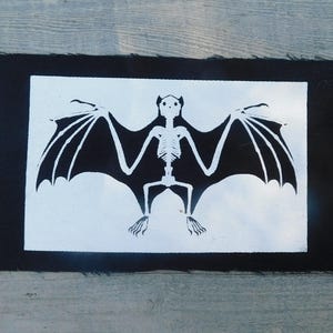 Op de afbeelding: Een zwart-wit vleermuisskeletontwerp op een rechthoekige patch. De vleermuis heeft uitgespreide vleugels en is gecentreerd op een witte achtergrond, omlijst door een zwarte rand. De patch is waarschijnlijk voor kleding of accessoires.