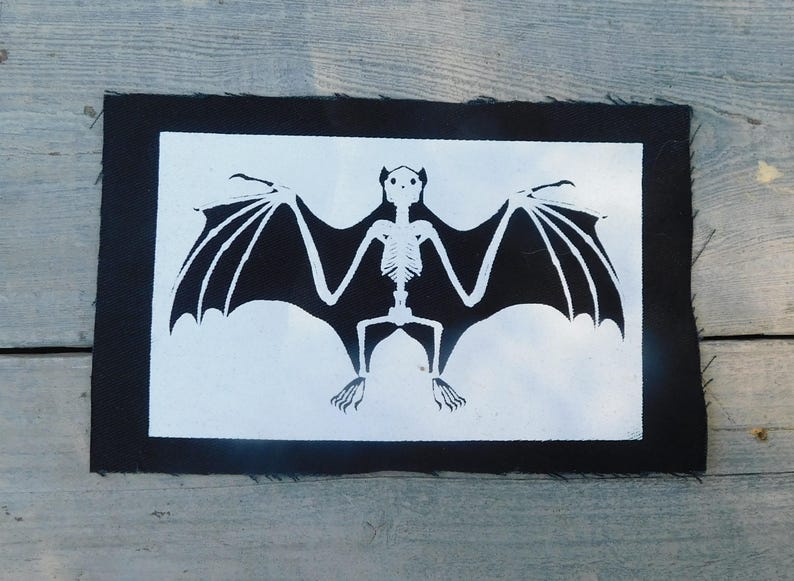 Op de afbeelding: Een zwart-wit vleermuisskeletontwerp op een rechthoekige patch. De vleermuis heeft uitgespreide vleugels en is gecentreerd op een witte achtergrond, omlijst door een zwarte rand. De patch is waarschijnlijk voor kleding of accessoires.
