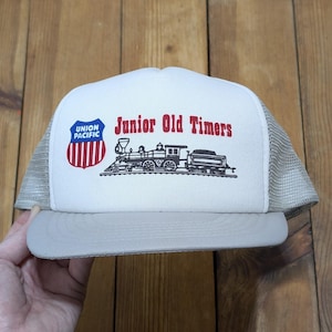May include: A white and tan trucker hat with the text "Junior Old Timers" in red. The hat features a Union Pacific logo and a black illustration of a train. The hat has a mesh back and a curved brim.