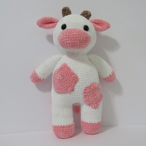 May include: A white crocheted cow plush toy with pink accents. The cow has a pink nose, pink ears, and pink spots on its body.