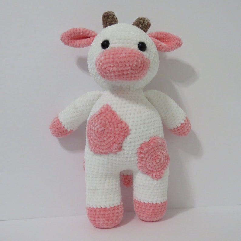 May include: A white crocheted cow plush toy with pink accents. The cow has a pink nose, pink ears, and pink spots on its body.