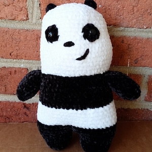 May include: A black and white crocheted panda plush toy with a black nose and black eyes. The panda is sitting on a brick surface.