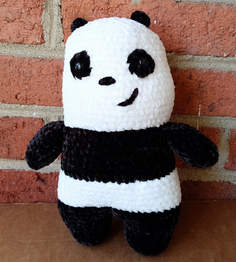 May include: A black and white crocheted panda plush toy with a black nose and black eyes. The panda is sitting on a brick surface.