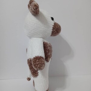 May include: A white crocheted plush toy cow with brown spots. The cow has a white face with brown ears and a brown nose. The cow is standing on a white surface.