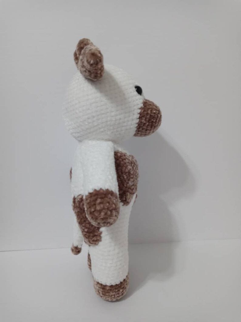 May include: A white crocheted plush toy cow with brown spots. The cow has a white face with brown ears and a brown nose. The cow is standing on a white surface.