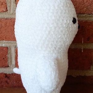 May include: White plush toy shaped like a bear, standing next to a brick wall. The bear has a small black eye and a textured surface.