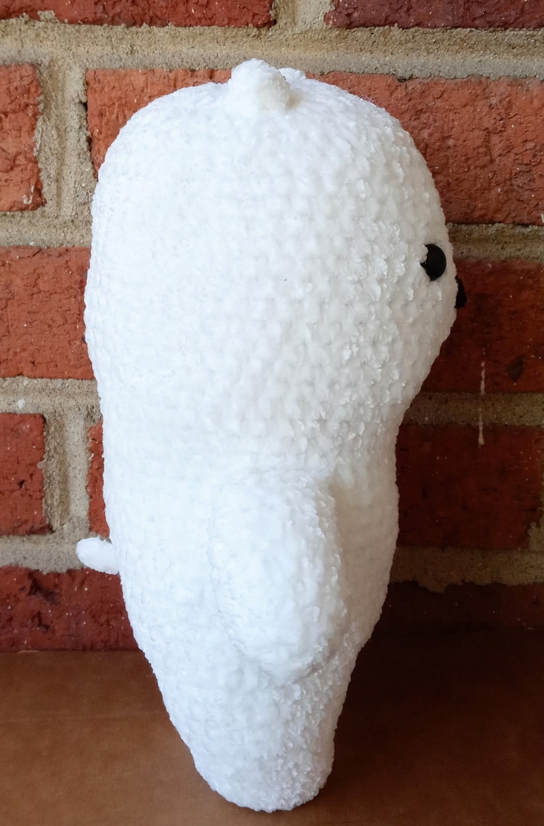 May include: White plush toy shaped like a bear, standing next to a brick wall. The bear has a small black eye and a textured surface.
