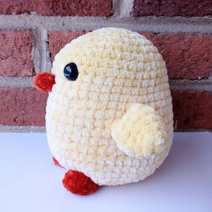 Puede incluir: Un juguete de pollito amarillo tejido a crochet con patas rojas y un ojo negro. El pollito est&aacute; sentado sobre una superficie blanca frente a una pared de ladrillo.