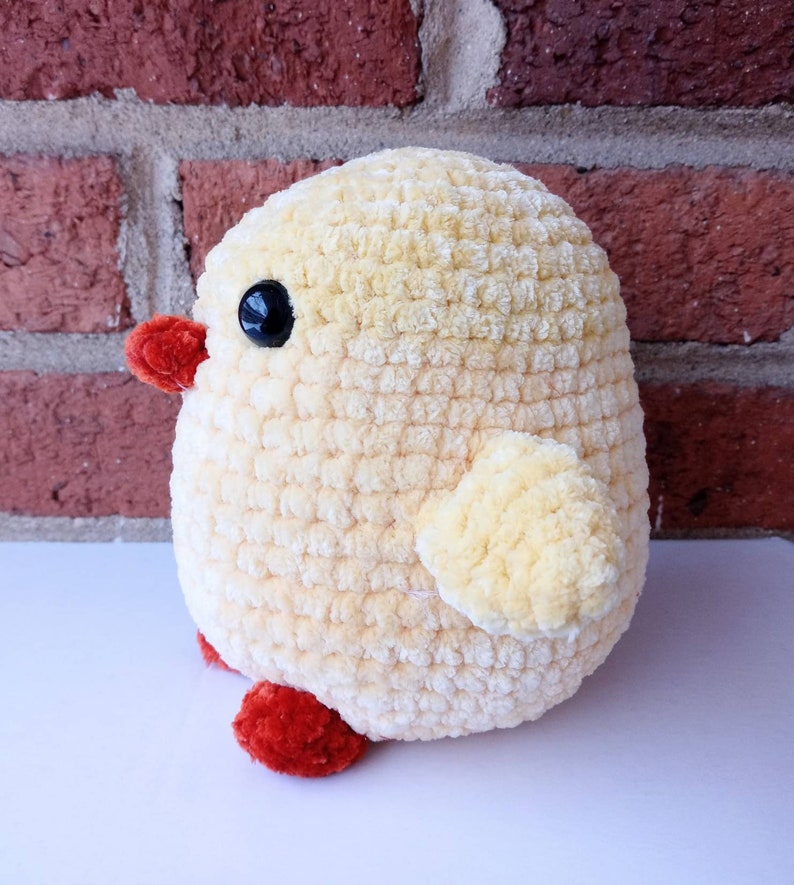Puede incluir: Un juguete de pollito amarillo tejido a crochet con patas rojas y un ojo negro. El pollito est&aacute; sentado sobre una superficie blanca frente a una pared de ladrillo.