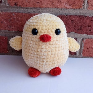 Puede incluir: Un juguete de peluche amarillo de pollito tejido a crochet con pies rojos y ojos negros. El pollito est&aacute; sentado sobre una superficie blanca.