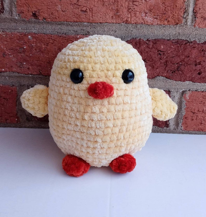 Puede incluir: Un juguete de peluche amarillo de pollito tejido a crochet con pies rojos y ojos negros. El pollito est&aacute; sentado sobre una superficie blanca.