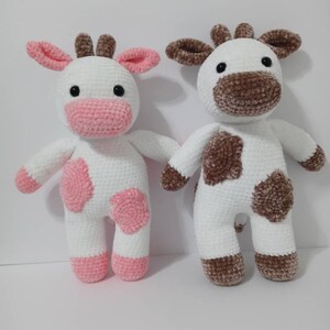 May include: Two crocheted cow plushies, one with pink spots and the other with brown spots. Both are white with black eyes and brown noses.