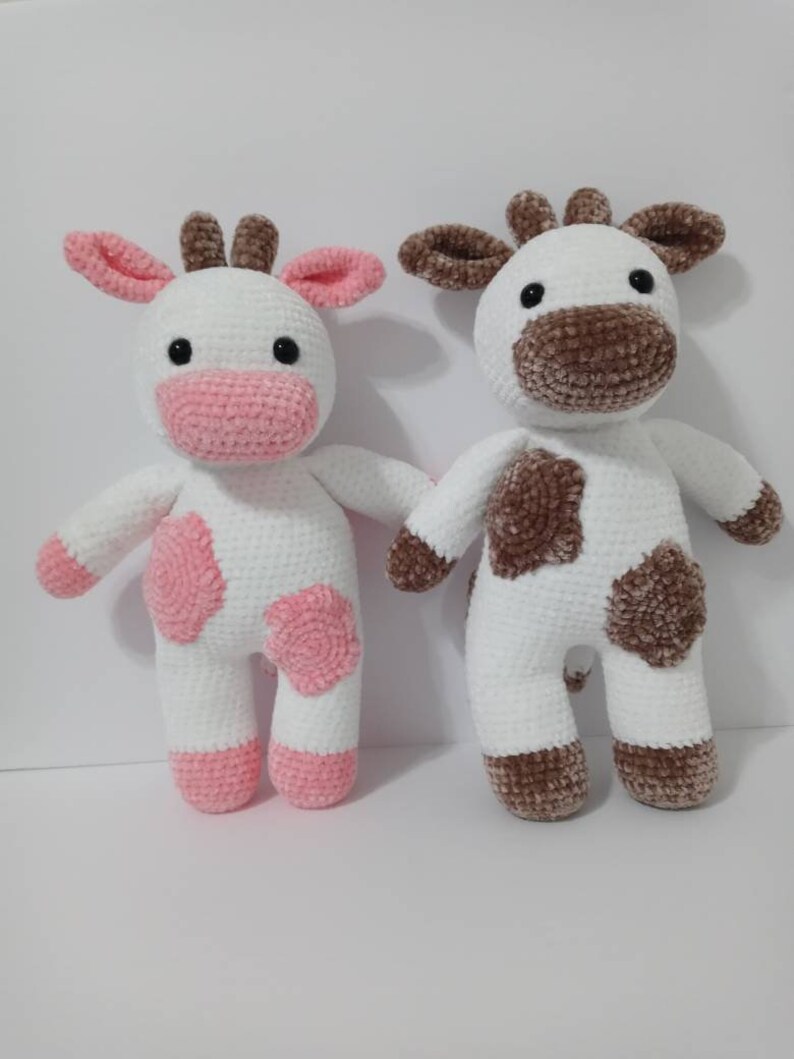 May include: Two crocheted cow plushies, one with pink spots and the other with brown spots. Both are white with black eyes and brown noses.