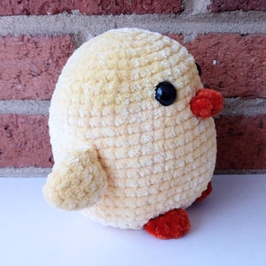 Puede incluir: Un juguete de peluche de pollito amarillo tejido a crochet con ojos negros y patas naranjas. El pollito est&aacute; sentado sobre una superficie blanca.