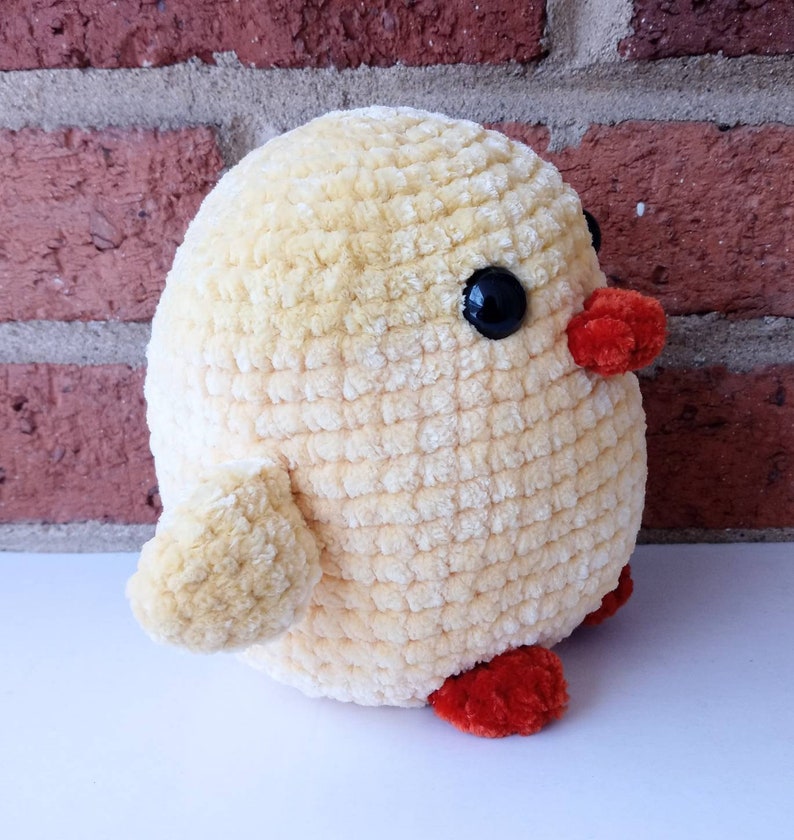 Puede incluir: Un juguete de peluche de pollito amarillo tejido a crochet con ojos negros y patas naranjas. El pollito est&aacute; sentado sobre una superficie blanca.