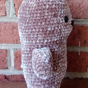May include: A plush crocheted teddy bear with a light brown colour and a velvety texture. The bear is standing on a brown surface.