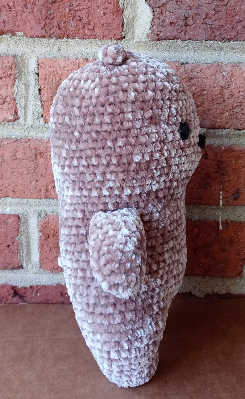 May include: A plush crocheted teddy bear with a light brown colour and a velvety texture. The bear is standing on a brown surface.