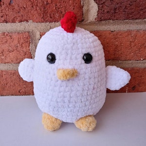 Puede incluir: Un juguete de peluche de pollo blanco tejido a crochet con ojos negros, un pico amarillo y una cresta roja en la cabeza.