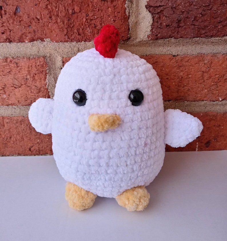 Puede incluir: Un juguete de peluche de pollo blanco tejido a crochet con ojos negros, un pico amarillo y una cresta roja en la cabeza.
