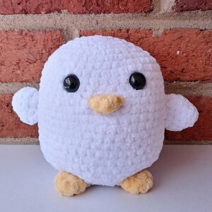 Puede incluir: Juguete de peluche de ping&uuml;ino blanco de crochet con ojos negros y pico naranja. El ping&uuml;ino est&aacute; de pie sobre una superficie blanca.