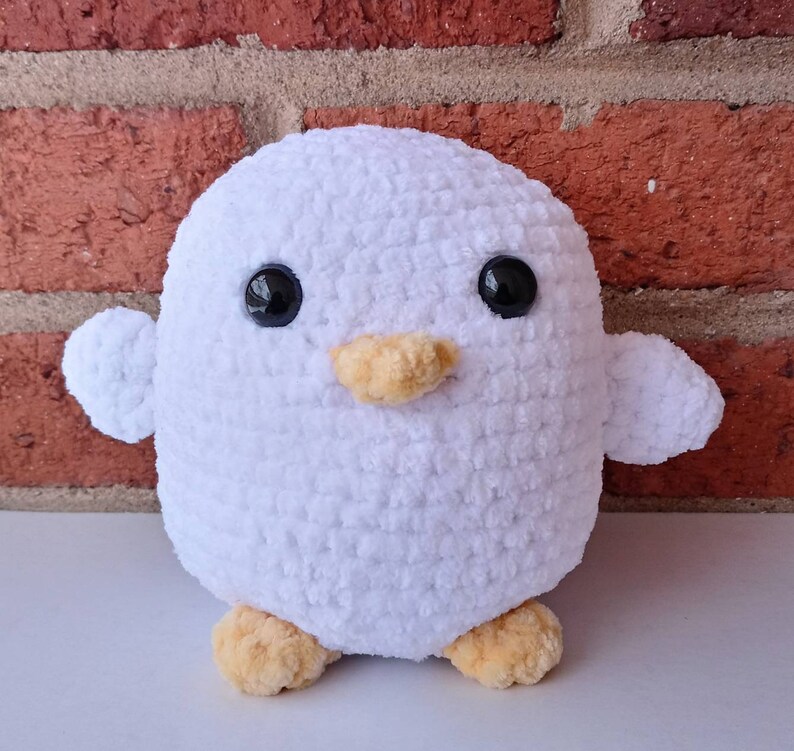 Puede incluir: Juguete de peluche de ping&uuml;ino blanco de crochet con ojos negros y pico naranja. El ping&uuml;ino est&aacute; de pie sobre una superficie blanca.