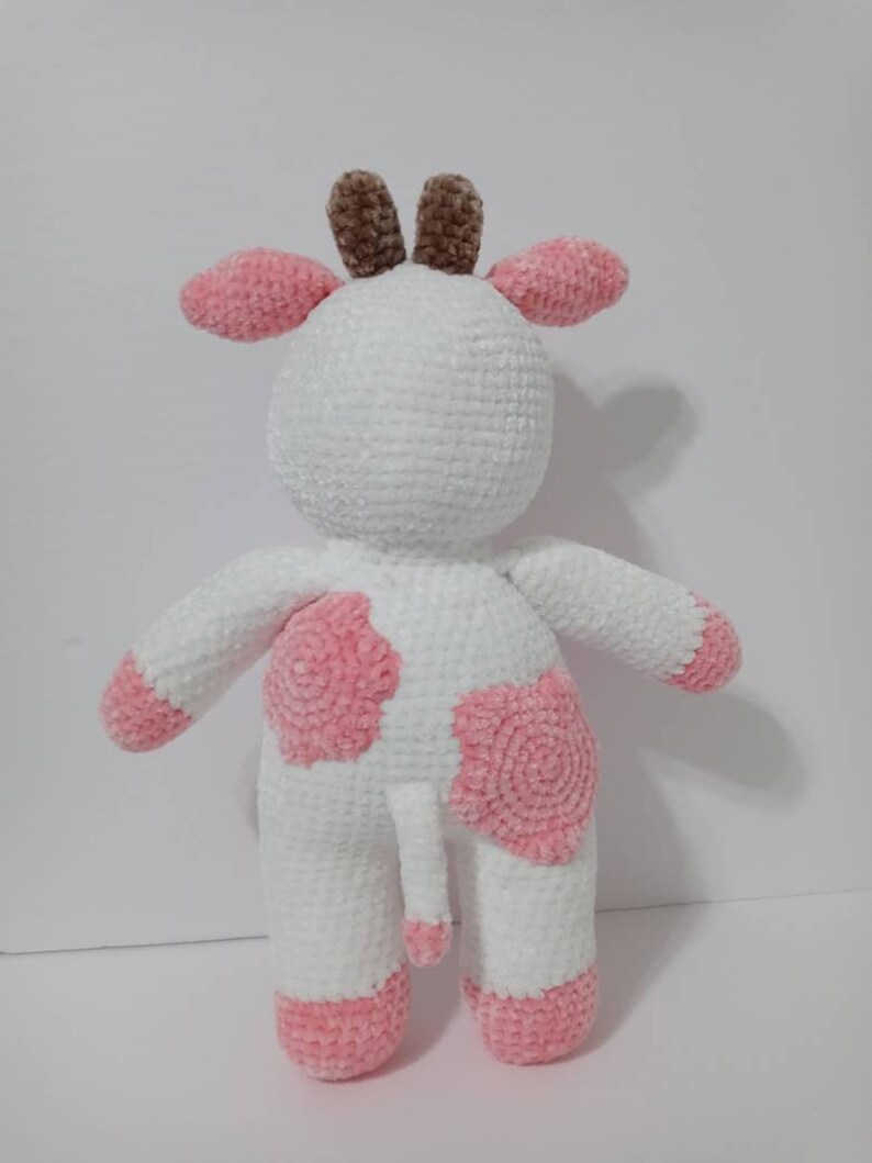 May include: A white crocheted cow plush toy with pink spots and brown horns. The toy has a pink tail.