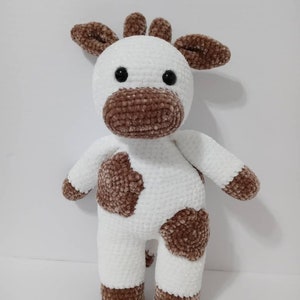 May include: A white crocheted stuffed animal with brown spots, resembling a cow. The animal has a brown nose, brown ears, and black eyes.