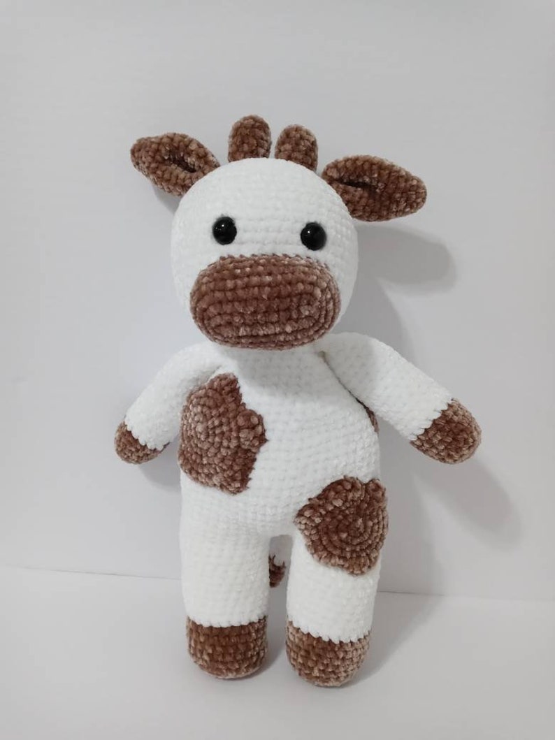 May include: A white crocheted stuffed animal with brown spots, resembling a cow. The animal has a brown nose, brown ears, and black eyes.