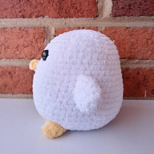 Puede incluir: Un juguete de peluche blanco de crochet con forma de p&aacute;jaro con ojos negros y patas naranjas. El juguete est&aacute; sentado sobre una superficie blanca.
