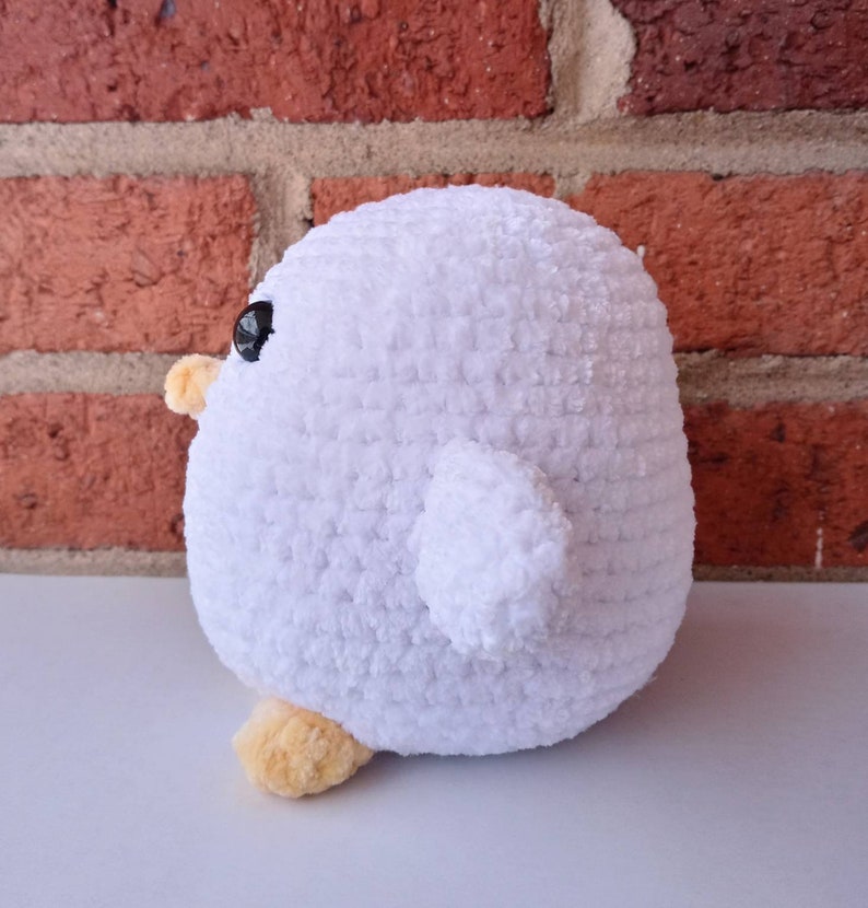 Puede incluir: Un juguete de peluche blanco de crochet con forma de p&aacute;jaro con ojos negros y patas naranjas. El juguete est&aacute; sentado sobre una superficie blanca.