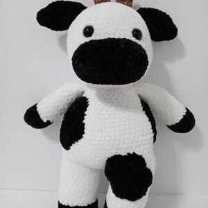May include: A white crocheted cow plush toy with black spots and brown horns. The toy has black eyes and a black nose.