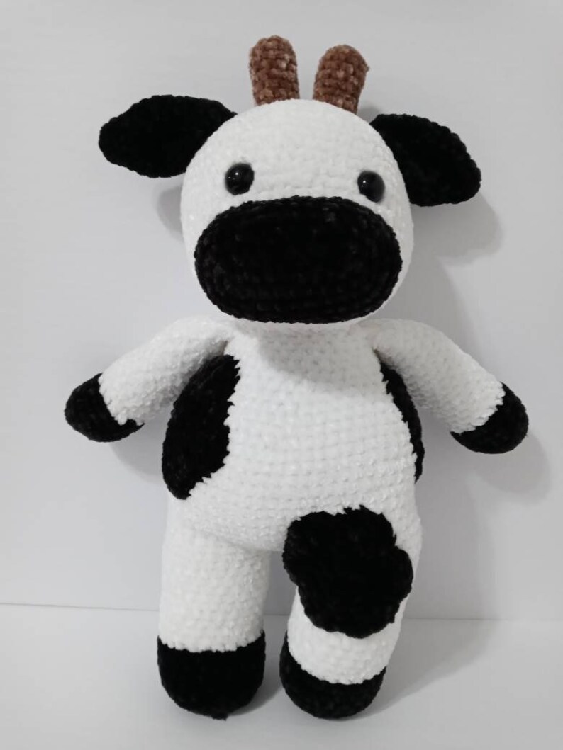 May include: A white crocheted cow plush toy with black spots and brown horns. The toy has black eyes and a black nose.