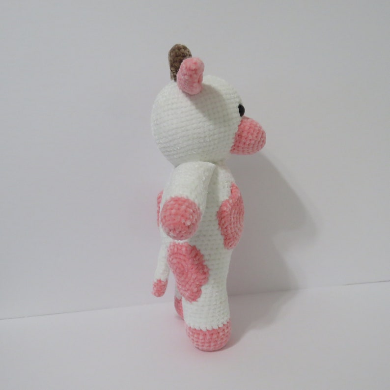 May include: A white crocheted cow plush toy with pink spots. The toy has a brown horn and black eyes.