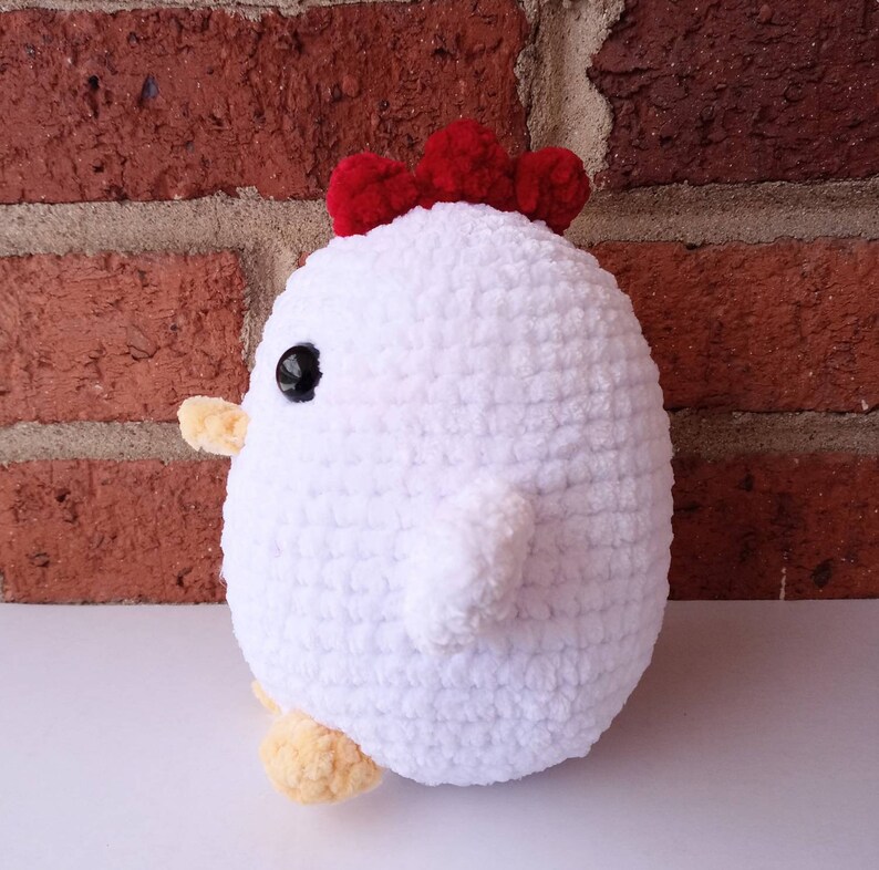 Puede incluir: Juguete de peluche de pollo blanco tejido a crochet con cresta roja y ojos negros. El juguete est&aacute; sentado sobre una superficie blanca.