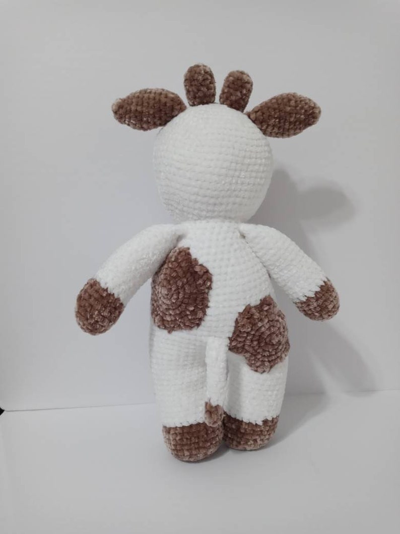 May include: A white crocheted plush toy cow with brown spots. The cow has a long tail and a brown crocheted head with ears.