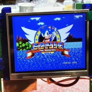 Könnte beinhalten: Nahaufnahme eines kleinen Bildschirms, der den Titelbildschirm des Videospiels "Sonic the Hedgehog" für Sega Genesis anzeigt. Der Bildschirm ist blau mit einem weißen und roten Banner, auf dem "Sonic the Hedgehog" und "SEGA" steht. Das Jahr 1991 ist in der rechten unteren Ecke des Bildschirms gedruckt.