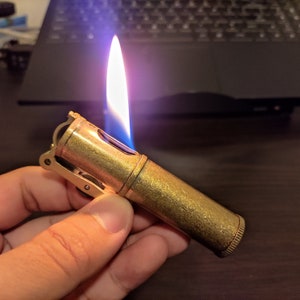 May include: A gold-colored lighter with a blue flame. The lighter has a textured surface and a small lever on the side.
