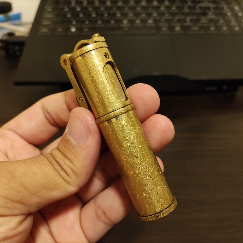 May include: A gold-colored metal lighter with a textured finish and a small window on the side. The lighter is held in a hand.