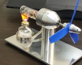 Op de afbeelding: Een kleine, zilveren, model stoommachine met een blauwe propeller. De motor wordt aangedreven door een klein vlammetje uit een glazen brandstoftank.