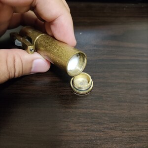 May include: A gold metal container with a hinged lid, open to reveal a white cotton swab inside. The container has a small, round, gold metal cap on the side.