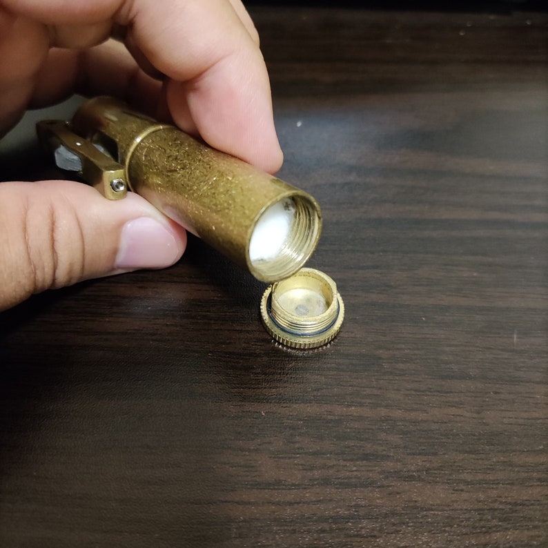 May include: A gold metal container with a hinged lid, open to reveal a white cotton swab inside. The container has a small, round, gold metal cap on the side.