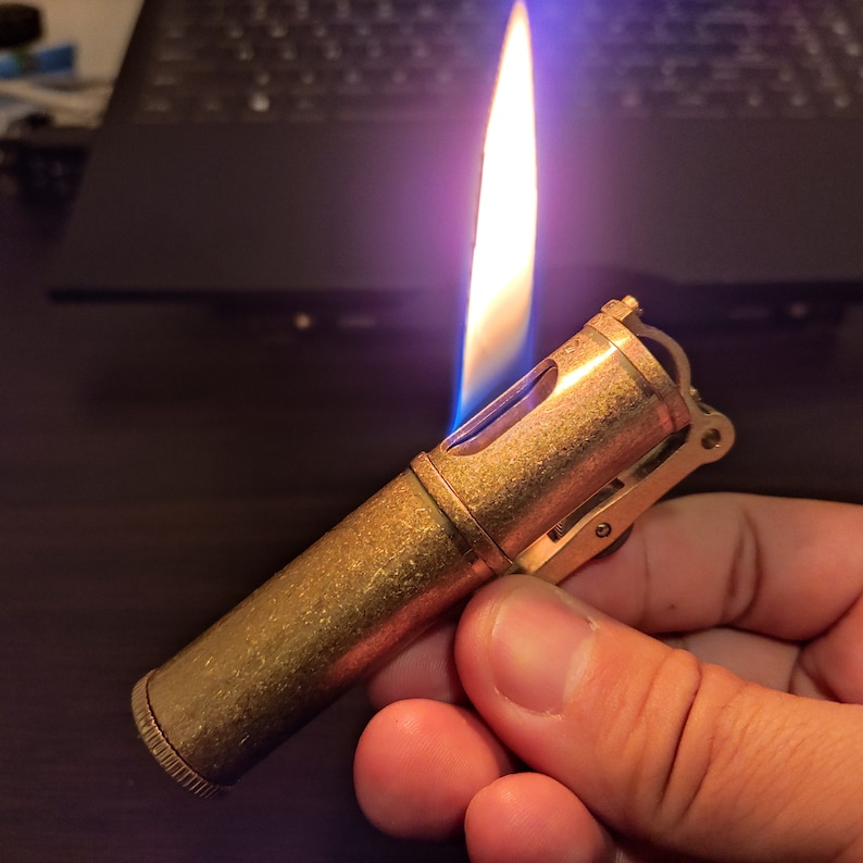 May include: A brass lighter with a blue flame. The lighter has a textured surface and a lever on the side.
