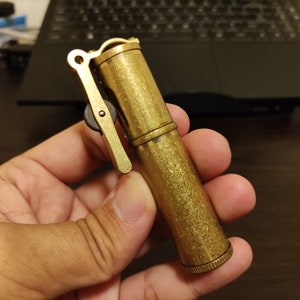 May include: A gold-colored metal lighter with a textured finish and a lever on the side. The lighter is being held in a hand.