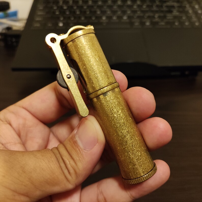 May include: A gold-colored metal lighter with a textured finish and a lever on the side. The lighter is being held in a hand.