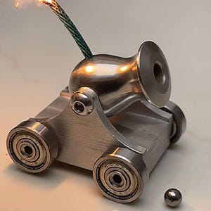 Peut inclure: Un canon miniature en métal argenté monté sur un chariot avec quatre roues à roulements à billes. Une mèche verte est insérée dans le haut du canon et une petite bille en métal repose sur la surface devant le canon.